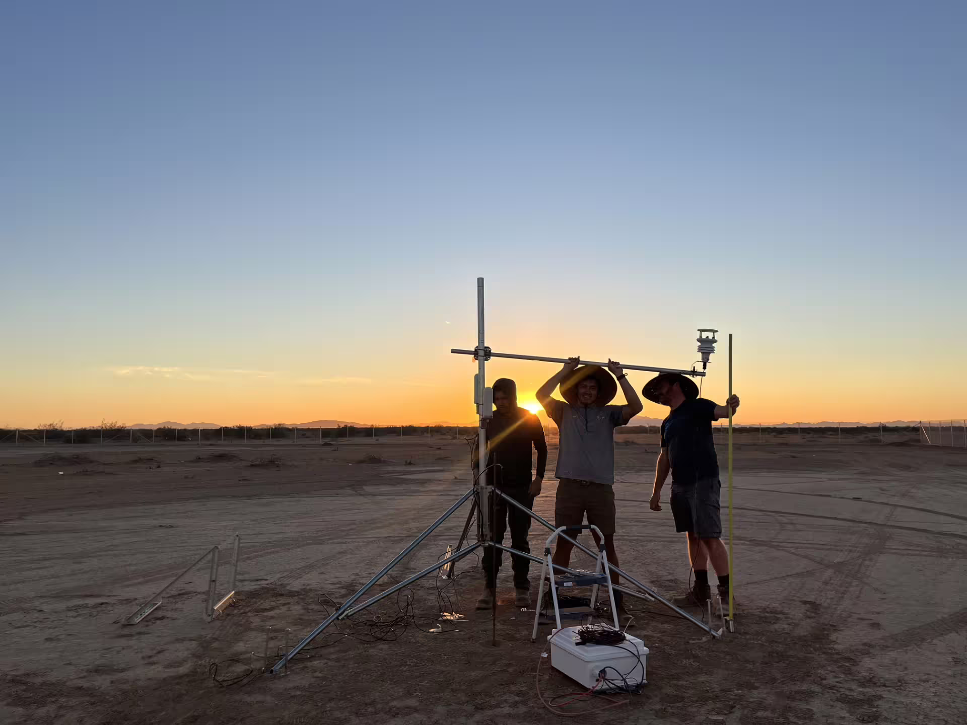 Three researchers set up meteorology equipment in a desert, backlit by setting sun